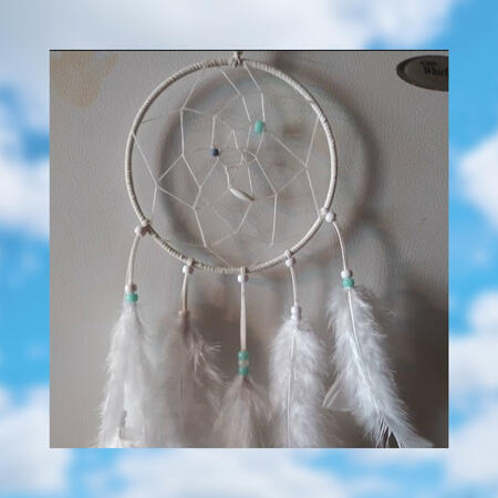 White dreamcatcher with white and blue beads, and white feathers. Blue sky with white clouds as background, blurred.